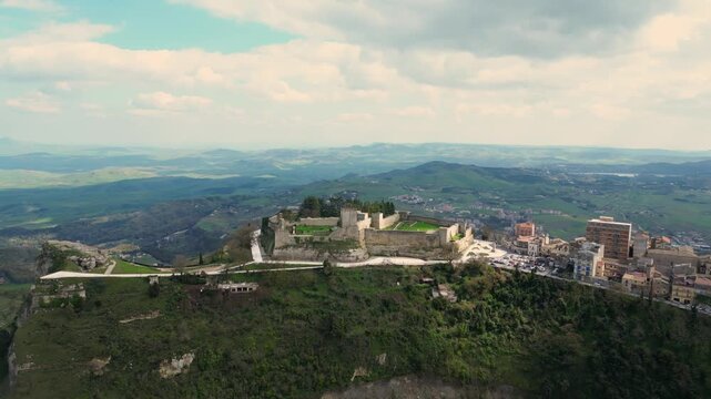 Aerial view of ancient castle ruins on a hilltop overlooking lush green valleys and distant mountains in Sicily, showcasing the historical architecture and natural beauty