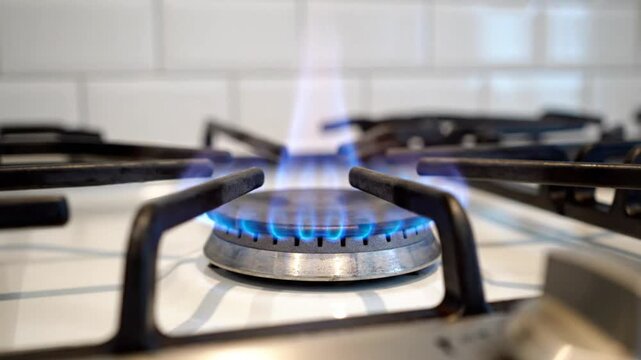 Close up of a gas stove burner with blue flames and a background of white tile in a kitchen