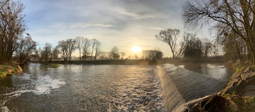 Morning sunrise over the Opava River with a small weir, captured in spring in the Czech Republic. The panoramic scene highlights soft golden light, calm water surface, and peaceful Czech countryside.
