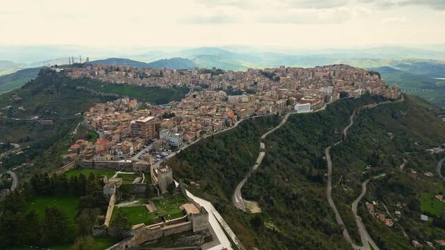 Aerial view of Enna, Sicily, showcasing the picturesque hillside town with winding roads, lush greenery, and historical architecture under a cloudy sky