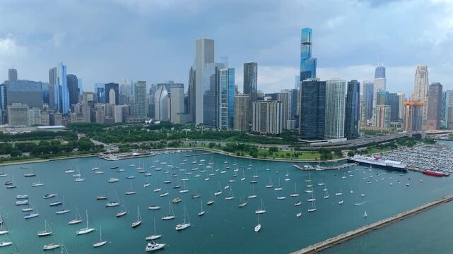 Drone footage captures Chicago downtown skyline with numerous sailboats anchored in Monroe Harbor. Modern skyscrapers rise behind Grant Park greenery under overcast summer sky.