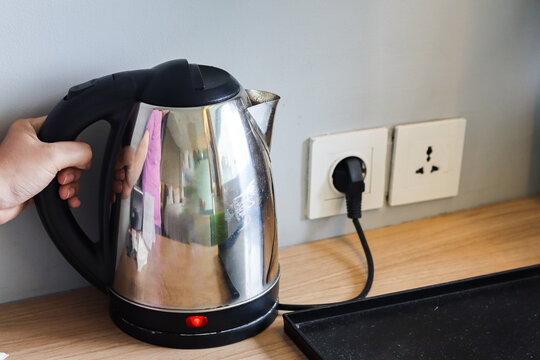 Electric kettle plugged into a wall outlet on a wooden table, with indicator light on, held by a woman's hand, used for boiling water in a kitchen or hotel room.