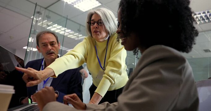Senior female manager mentoring her diverse team, pointing at a laptop screen while discussing a project in a contemporary glass walled office, fostering teamwork and professional development