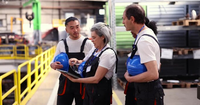 Multiethnic group of industrial workers discussing a project in a factory warehouse with a female supervisor holding a tablet, fostering collaboration and planning for production