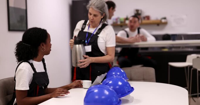 Two diverse female factory workers wearing overalls enjoy a coffee break together, with one woman pouring a hot beverage from a thermos for her colleague in the company canteen