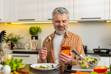 Middle-aged man enjoying salad while checking smartphone for calorie tracking weight loss plans in...