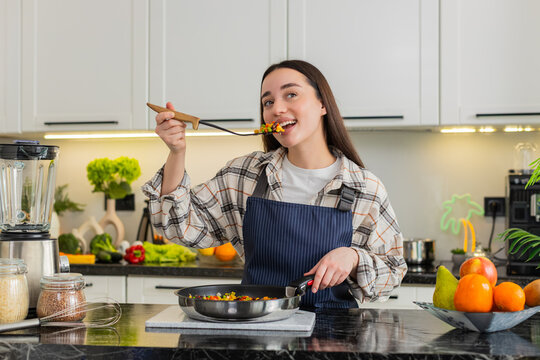 Young woman samples vegetables from frying pan while cooking in home kitchen for diet plan and daily wellness. Girl looks satisfied after taste test ready to serve homemade lunch in apartment indoors.