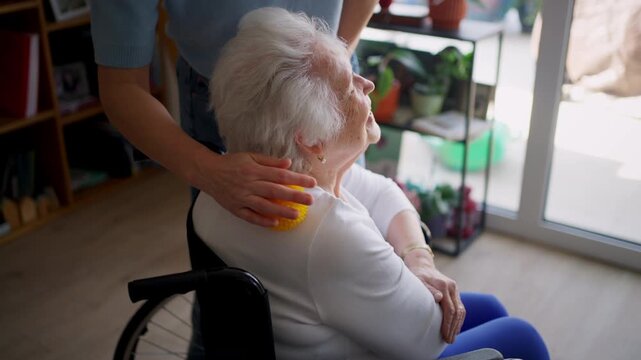 Daughter helping elderly mother in wheelchair with physiotherapy exercise at home.