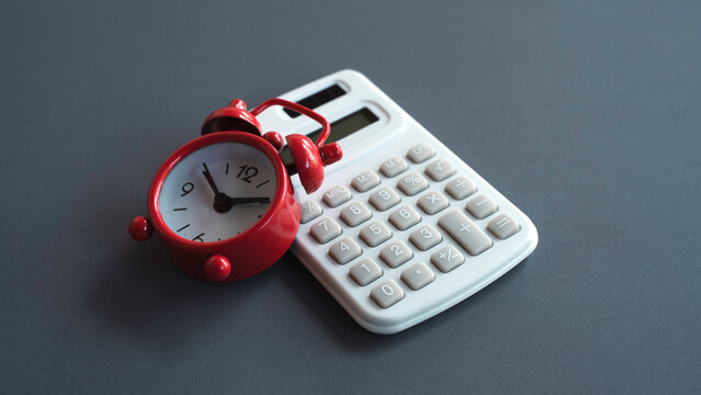 Red alarm clock resting on a white calculator on a dark surface representing financial deadlines, time value of money, and tax season