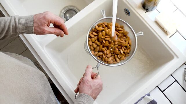 Man washes almonds in a metal colander under running water in a kitchen sink. Symbolizes healthy eating, clean food preparation, and mindful nutrition. Ideal for cooking, wellness, and dietary content