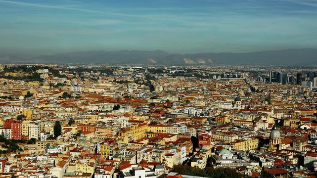 Naples dense cityscape stretches across the horizon during daylight. Historical architecture and crowded residential rooftops fill the urban panorama against a distant mountain backdrop.