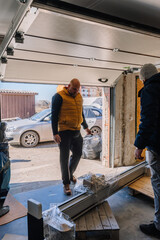 Fototapeta premium Man in yellow vest inspects delivery items inside a garage with an open door, revealing a car parked outside and a sunny day visible
