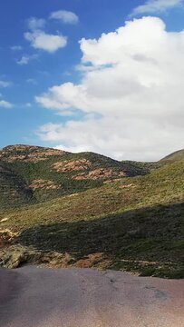 Scenic view of rugged mountains and green hills under a blue sky with clouds in the Delta du Fango region of Corsica. Natural terrain and dramatic light create a tranquil atmosphere.