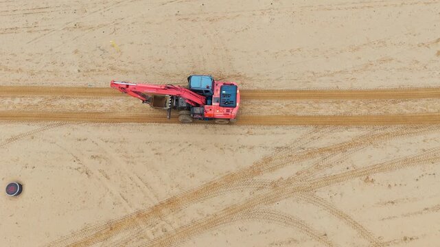 Aerial view of red excavator on sandy terrain. Machine moves slowly across loose earth. No people visible, no animals or plants present. Ideal for construction, mining, or land development projects