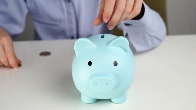 Hand inserts coin into piggy bank. Piggy bank is light blue, round, with simple eyes. Person wears light blue shirt, sits at table. Scene suggests saving money or financial planning