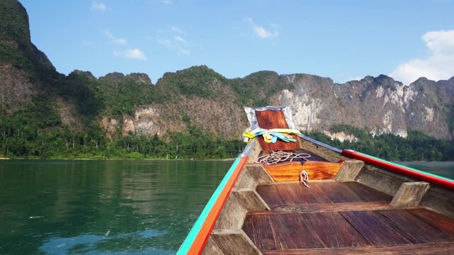 Longtail boat glides across calm, green waters. Mountains rise behind, covered in dense jungle. Sunlight highlights the boat's wooden deck. No people are visible, so no emotions can be inferred