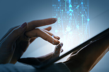 Close-up hands interacting with digital tablet showing flowing binary data and arrows on blue tech...