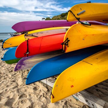 A colorful stack of kayaks on a sandy beach under a bright blue sky. The kayaks vary in color and are positioned with their hulls upward