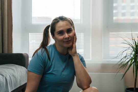 Smiling Caucasian Woman Seated By Window, Casual Blue Tee, Pigtails, Hand On Cheek, Soft Natural Light, Modern Apartment Interior With Plant And Bed,