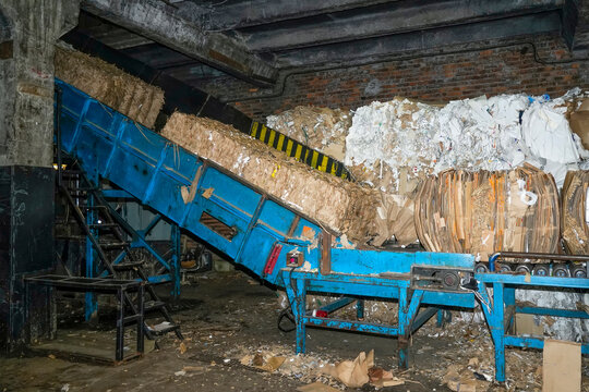 Blue conveyor belt moves bundles of recycled cardboard and paper up a slope at a recycling plant. Piles of compressed materials are visible in the dimly lit interior of the factory