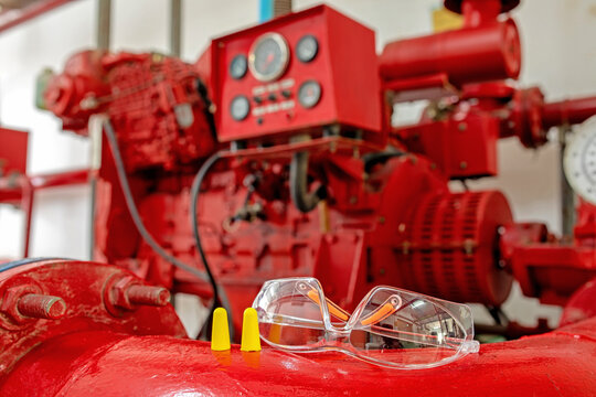 Protective clear eyewear and yellow earplugs resting on a red pipe in front of an autonomous fire fighting pump engine in a residential complex