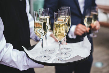 A waiter carries a tray of sparkling champagne flutes at a formal outdoor event. Guests in elegant suits stand in the blurred background, creating a sophisticated atmosphere for a luxury party. © Francis