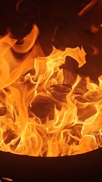 A close-up video of yellow and white ritual paper offerings, including joss paper, being consumed by bright orange flames inside a metal incinerator barrel. 