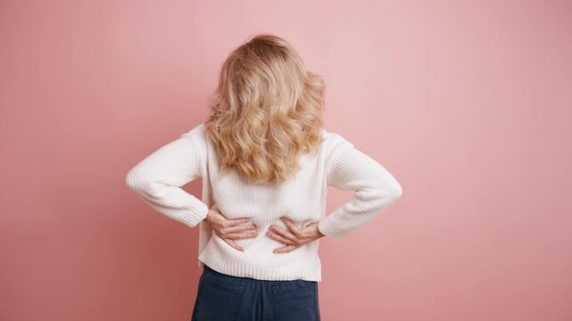 Rear view of a mature blonde woman massaging her lower back, experiencing sudden pain. Unhealthy female feeling muscle strain on a pink background