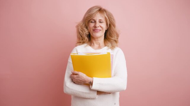 Cheerful mature woman holding a yellow document folder and smiling at the camera. An elegant business lady standing in a pink studio