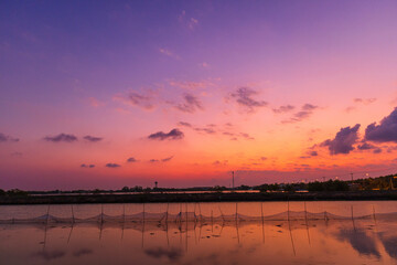 Obraz premium Serene Sunset Over Coastal Fish Pond with Purple Sky Reflection,A breathtaking sunset over a rural aquaculture farm, featuring a vibrant gradient sky from deep purple to fiery orange. The calm water.