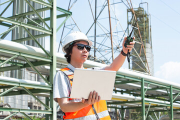 Worker Inspects Industrial Piping System with Laptop and Radio at Oil Refinery Site, Reviewing...
