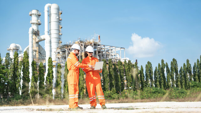 Two workers in orange coveralls and white helmets examine laptop alongside large industrial refinery plant in grassy setting, reviewing equipment data and monitoring operations, assessing plant safety