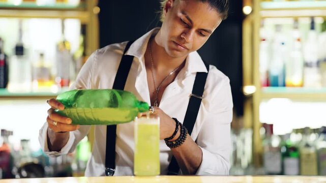 Concentrated male bartender in a white shirt and suspenders pouring a green beverage from a bottle into a tall glass at the bar counter