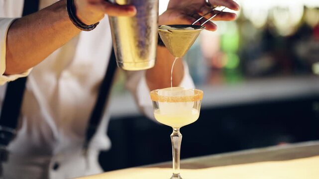 Close up of a skilled bartender pouring a freshly mixed cocktail from a shaker through a strainer into an elegant glass at a bar counter