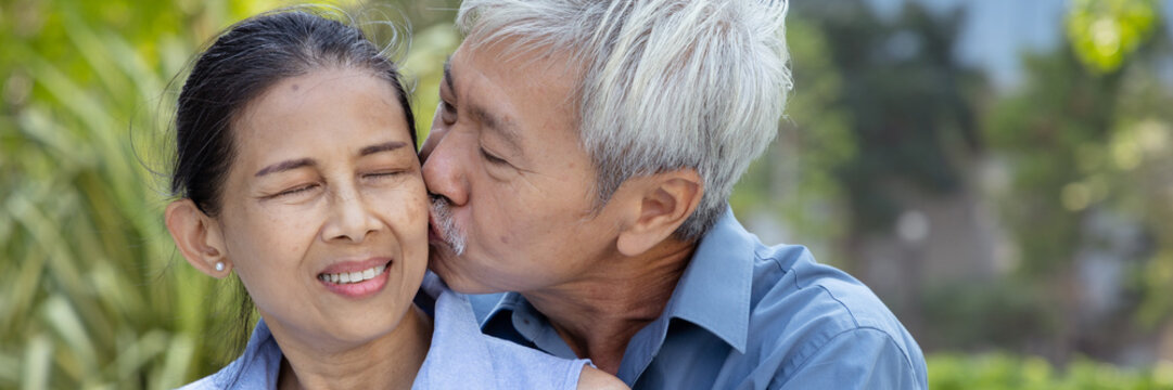 Happy smiling lovely Asian senior couple, elderly husband hugging and kissing on the cheek of his wife in urban park, concept image for old aged lovers togetherness, real human, real people model