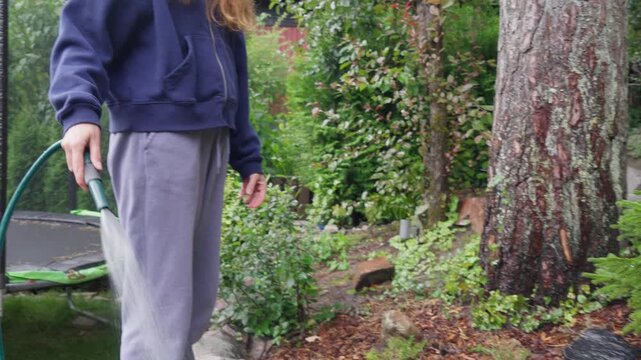 Young woman watering garden plants with a hose in backyard. Authentic domestic lifestyle, sustainable gardening and home maintenance concept in soft daylight.
