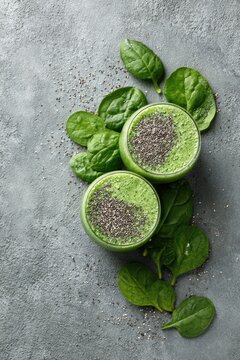 Two refreshing green smoothies topped with chia seeds and surrounded by fresh spinach leaves on a textured gray background.