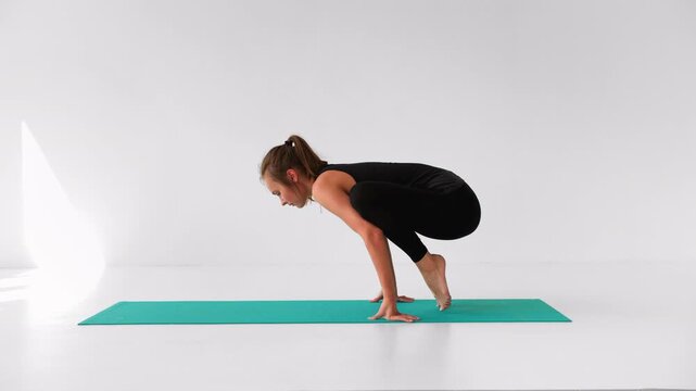 Young woman practicing the bakasana yoga pose on a turquoise mat in a white studio. Strength, balance, and flexibility training