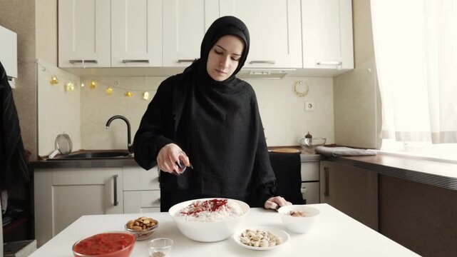 muslim woman in black hijab sprinkles seasoning over a large bowl of white rice in a cozy kitchen, documenting the hands-on preparation of a homemade festive family meal