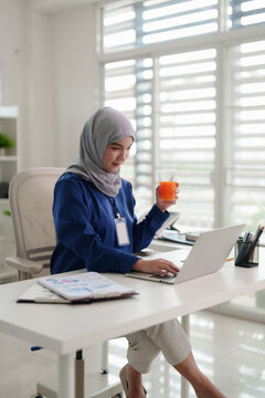 Asian Muslim adult woman working laptop corporate business tasks modern bright office environment focus healthy beverage professional development strategy