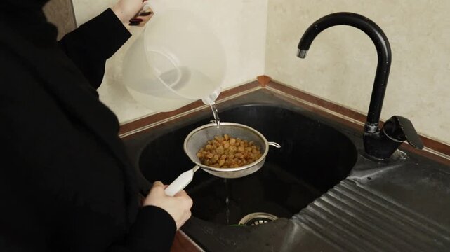 close-up of water being poured over golden raisins in a metal sieve above a dark kitchen sink, capturing a careful rinsing step in festive rice dish preparation