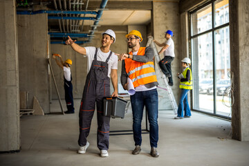 Fototapeta premium Construction workers with a toolbox and blueprints gesturing at a building site.