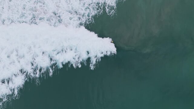 Top-down aerial view of a surfer riding a yellow surfboard on a breaking wave in clear green ocean water.