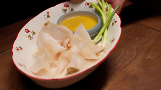 Thinly sliced cured fatback arranged on a decorative plate with mustard dipping sauce and fresh green onions served on a wooden table in warm lighting