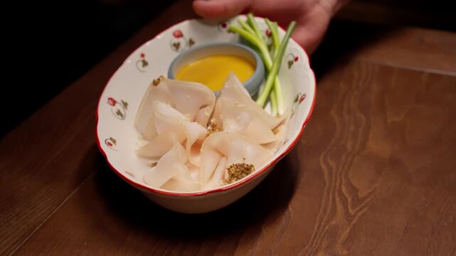 Delicate thinly sliced white fish appetizer with sesame garnish, green onion stalks, and smooth yellow dipping sauce served in a decorative oval dish on wooden table
