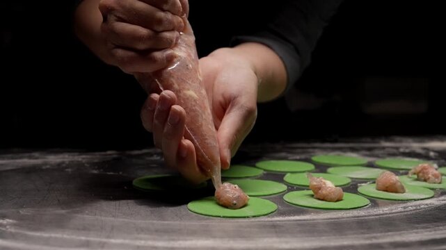 Closeup hands piping savory meat filling onto vibrant green dough circles on floured surface, preparing handmade dumplings in low light kitchen setting