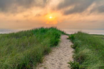 A winding sandy path cuts through lush green dune grass, leading the eye toward a vast, calm ocean. Above the horizon, a glowing sun breaks through a heavy layer of golden and grey clouds at sunset.