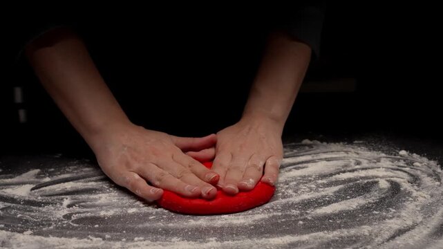 Hands shaping bright red dough on a floured dark surface, gently pressing and preparing creative homemade pastry under dramatic low key kitchen lighting