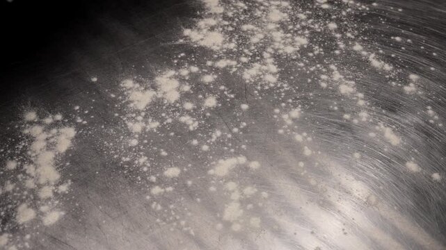 Fine white powder scattered across a scratched metallic surface, showing subtle textures, light reflections, and abstract patterns forming between the particles