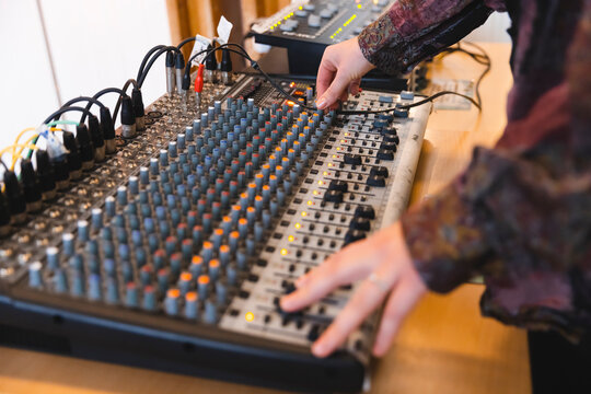 Close-up of a person's hands operating a professional analog audio mixing console with cables, adjusting knobs and faders in a recording studio or live sound environment for music production.